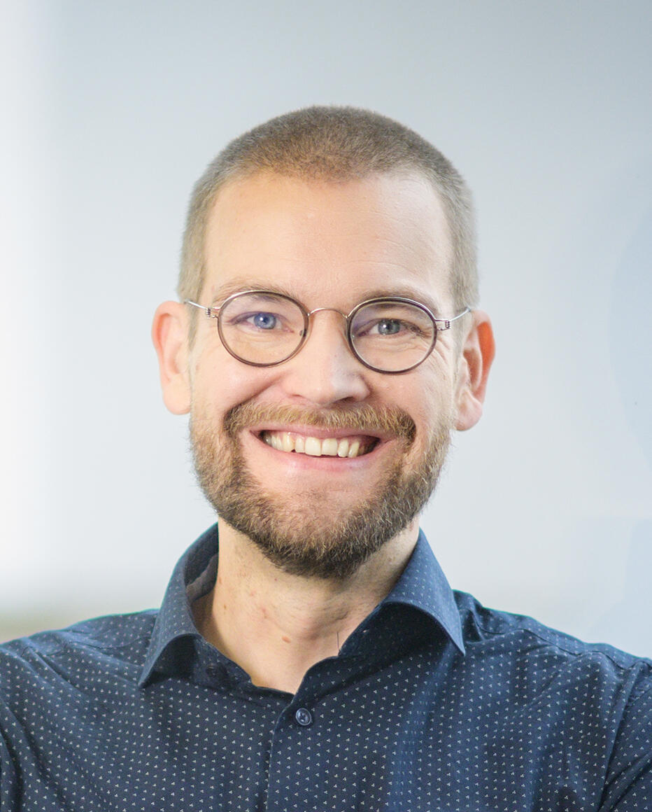 Hymyilevä mies, jolla on silmälasit, parta ja lyhyeksi leikattu tukka. Hänellä on tummansininen kauluspaita, ja taustana on vaalea seinä.